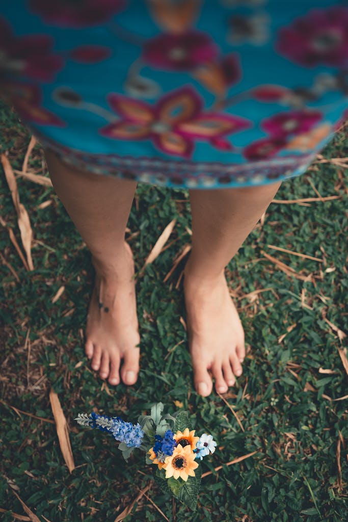 A barefoot woman standing in a floral dress on green grass with wildflowers at her feet.